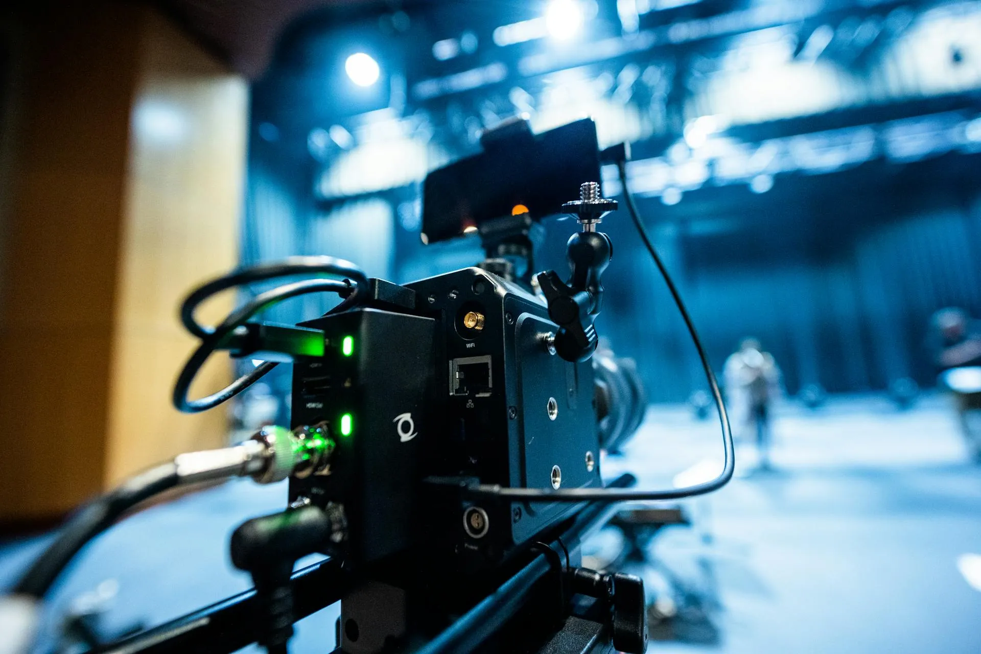 A television camera on set with a blue-lit backdrop and crew standing by